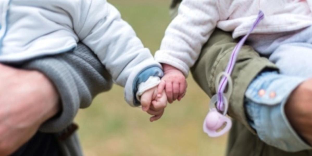 Two adults, heads not shown, holding two infants, heads not shown. Infants are holding hands.